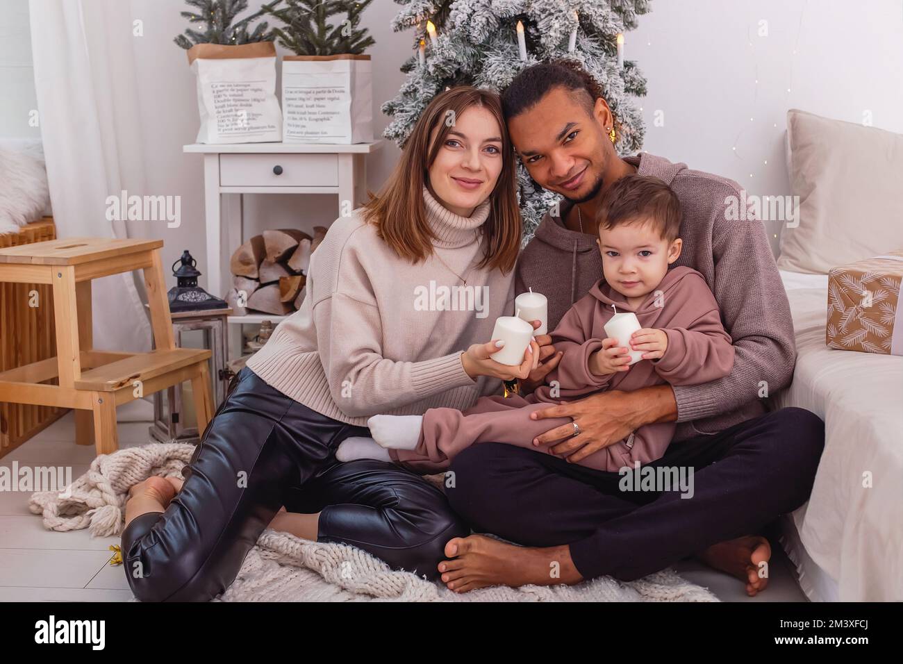 Una famiglia giovane e varia con un bambino è seduta con la candela vicino ad un albero di Natale Foto Stock