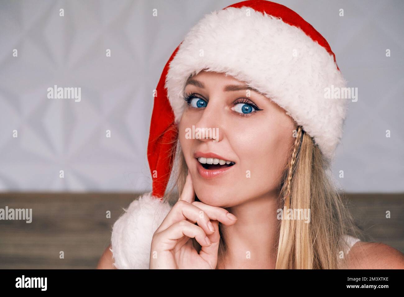 Santa ragazza sorridente e sognante che indossa un cappello rosso che tocca la guancia con un dito guardando la macchina fotografica. Ritratto di bella giovane donna con nel cappello di Babbo Natale Foto Stock