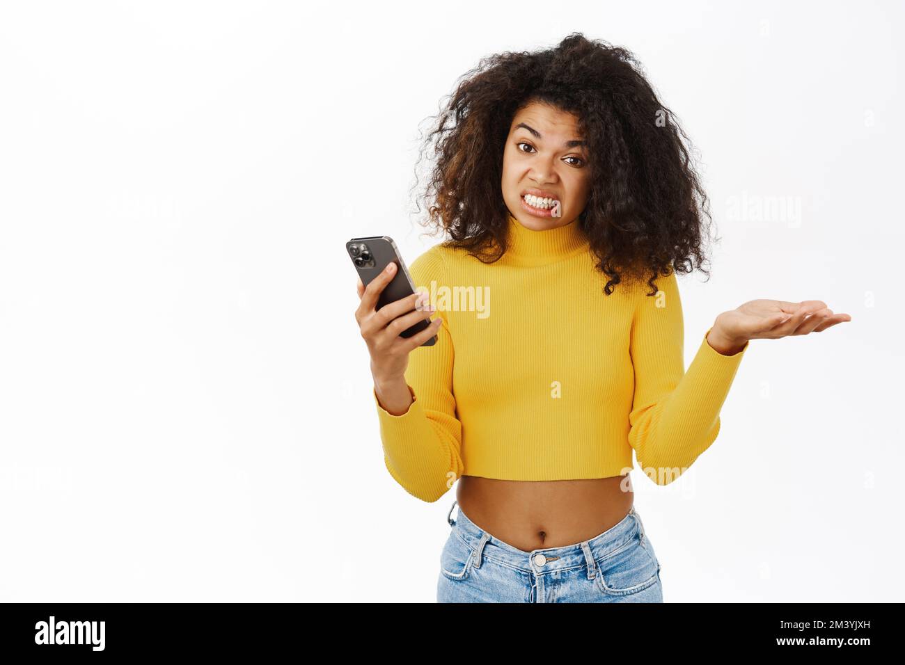 Ragazza afroamericana arrabbiata, in cerca di frustrazione, tenendo smartphone con l'espressione del volto delusa, smth odio, in piedi su sfondo bianco Foto Stock