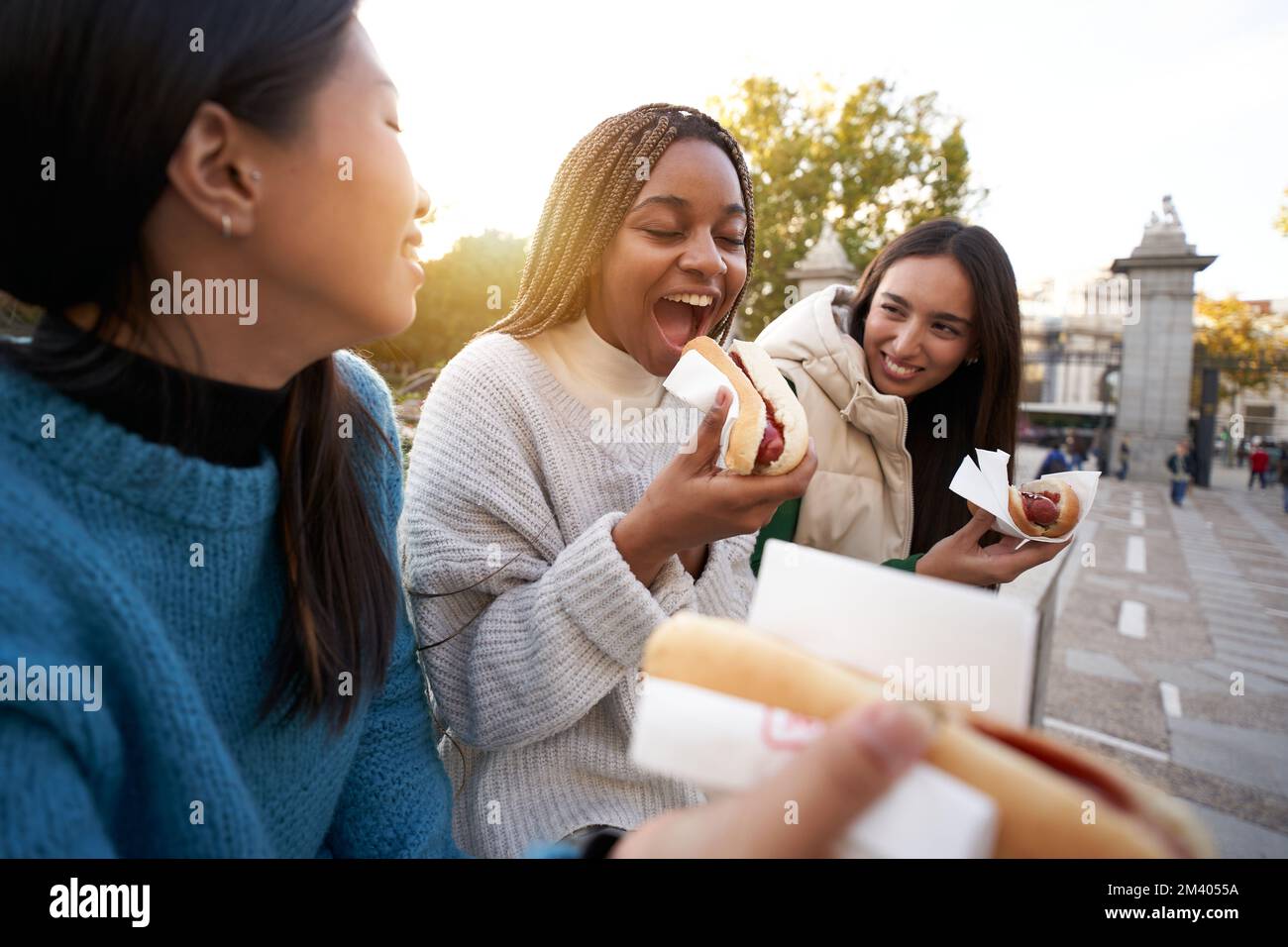 Ragazze allegre mangiare cibo di strada take away. Tre amici di Girlfriends felici che prendono i cani caldi nel parco Foto Stock