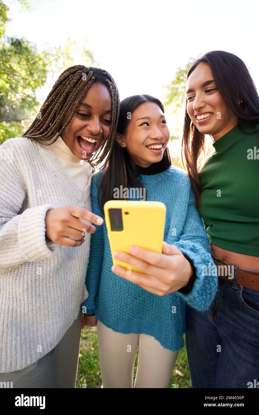 Verticale. Tre giovani donne che usano il telefono mentre si divertono nel parco e ridono. App per social media Foto Stock