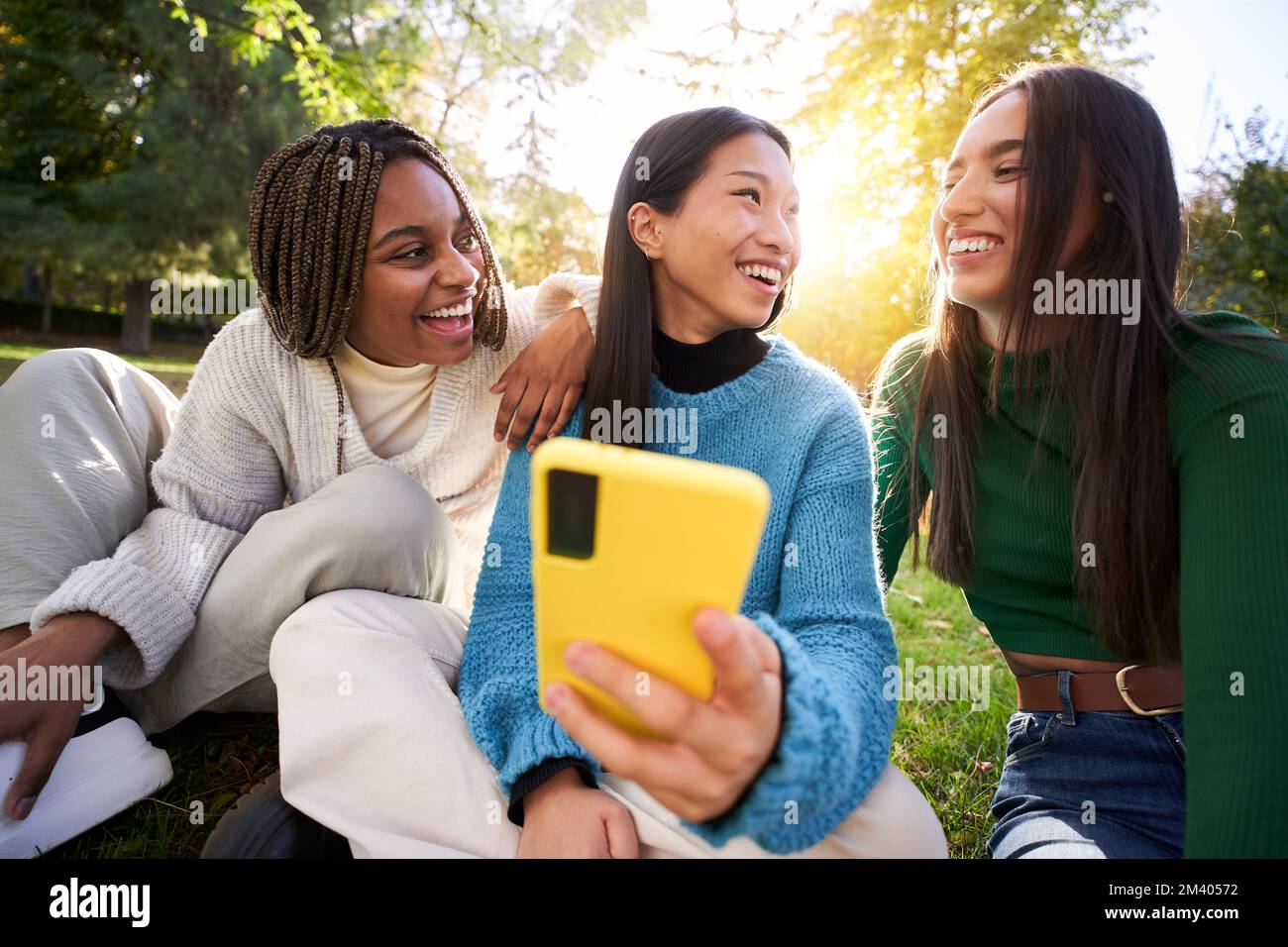 Tre giovani donne che usano una cella mobile mentre si divertono nel parco e ridono. App per social media Foto Stock