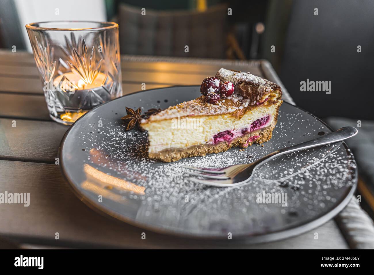 Fetta di cheesecake con ciliegie servita su piatto grigio con zucchero a velo e stelle di anice. Cottura di Natale. Dessert festivo. A lume di candela. Scatto orizzontale in interni. Foto di alta qualità Foto Stock