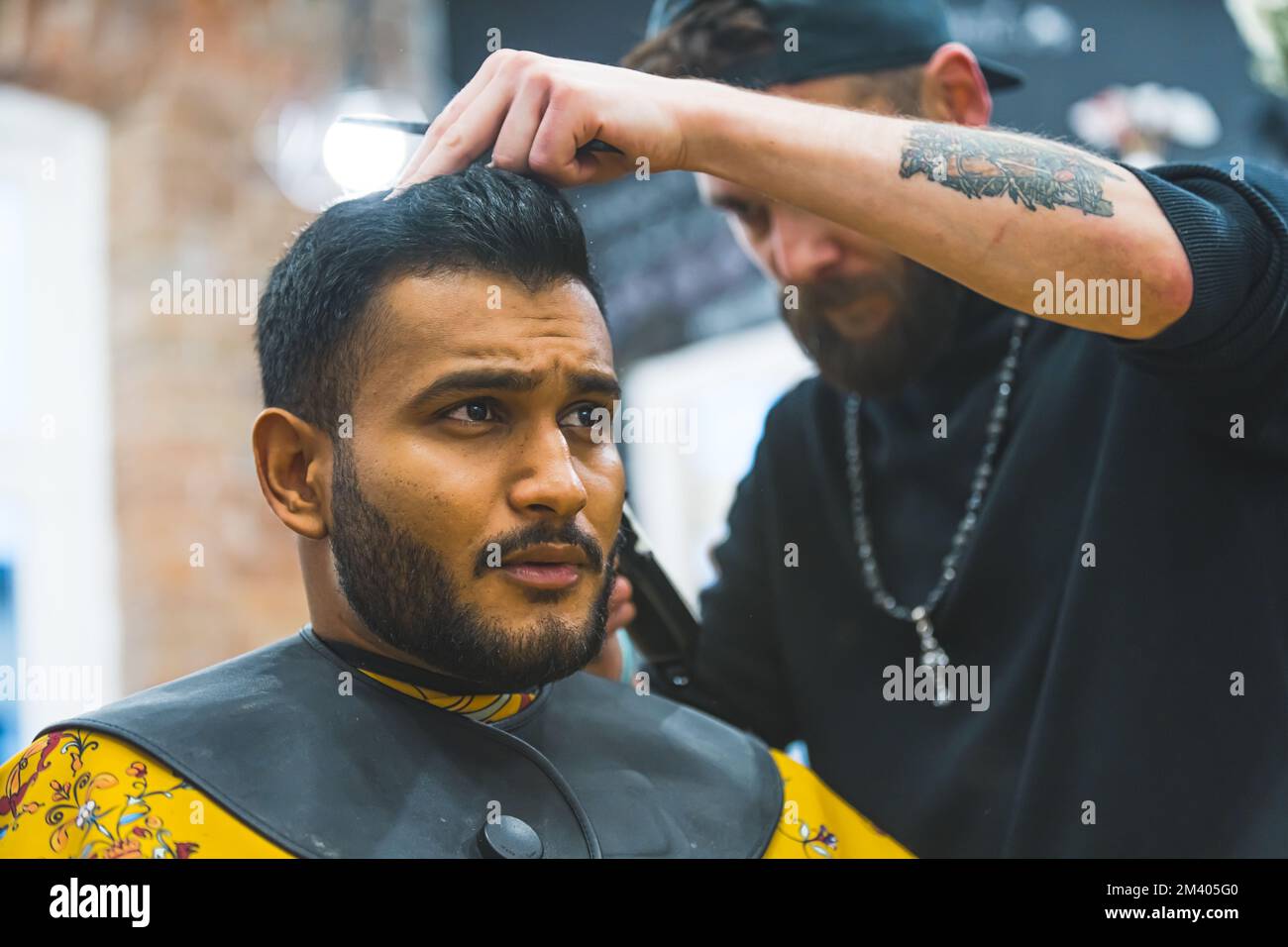 Barbiere professionale styling i capelli del suo cliente indiano felice in mantello giallo per un taglio di capelli in un barbiere. Foto di alta qualità Foto Stock