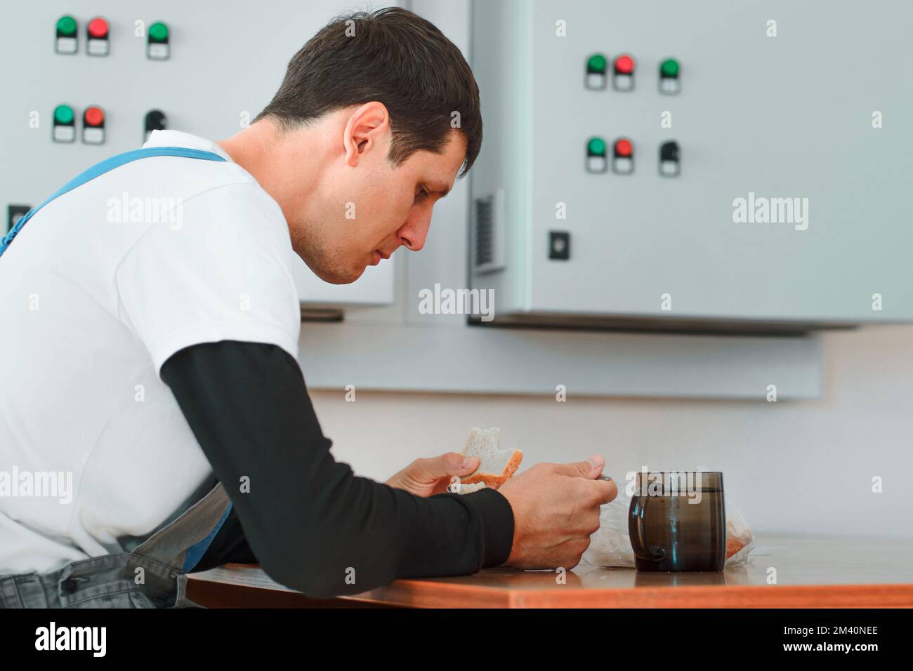Pausa pranzo. Mangiare sul posto di lavoro durante il lavoro. L'uomo caucasico in tuta da lavoro siede al tavolo nella sala di produzione e mangia dal contenitore. Il lavoratore ha pranzo in sala pausa. Foto Stock