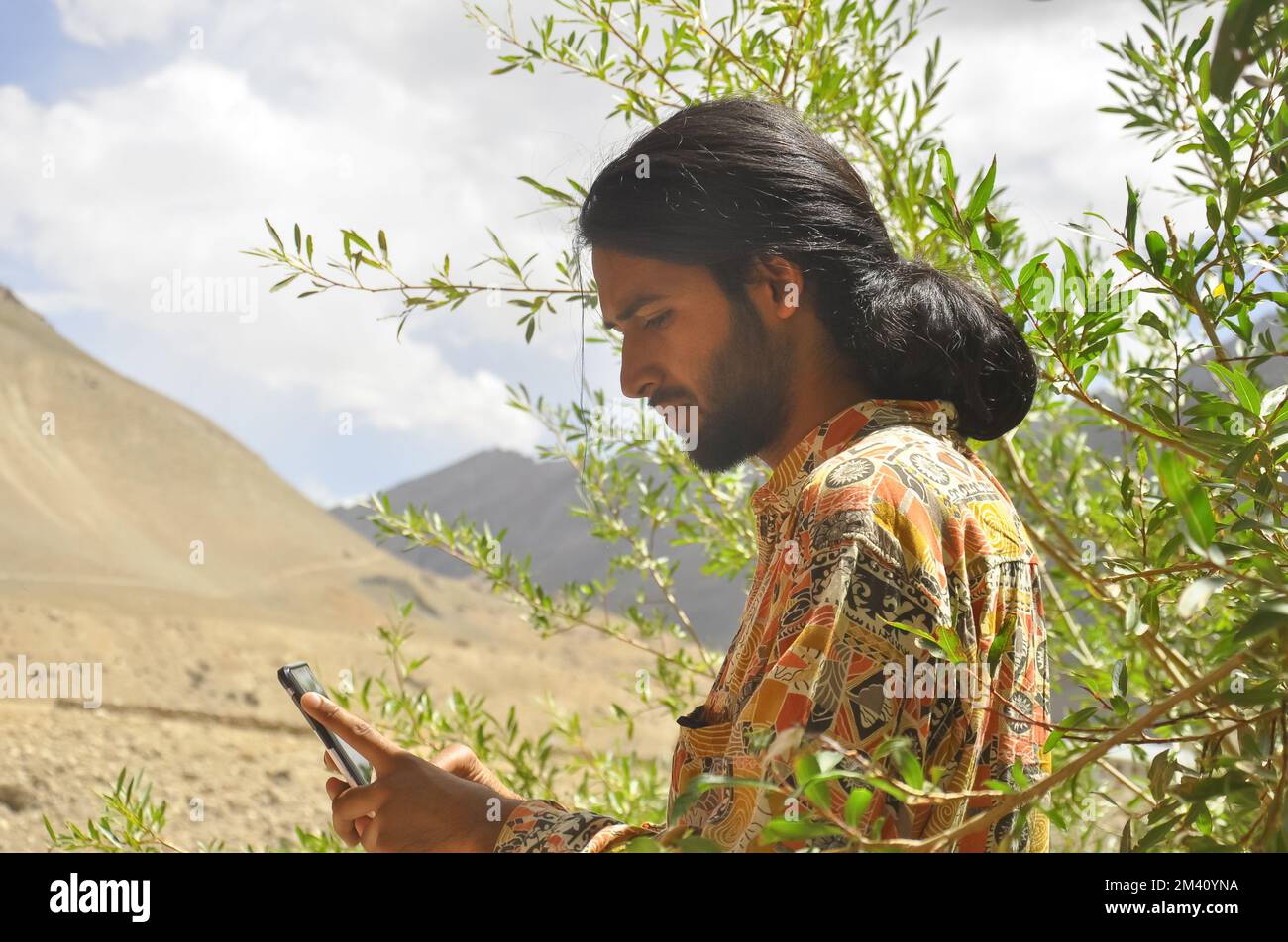 Vista laterale di un giovane con capelli lunghi e barba utilizzando il telefono con lo sfondo delle montagne della valle di Zanskar. Foto Stock