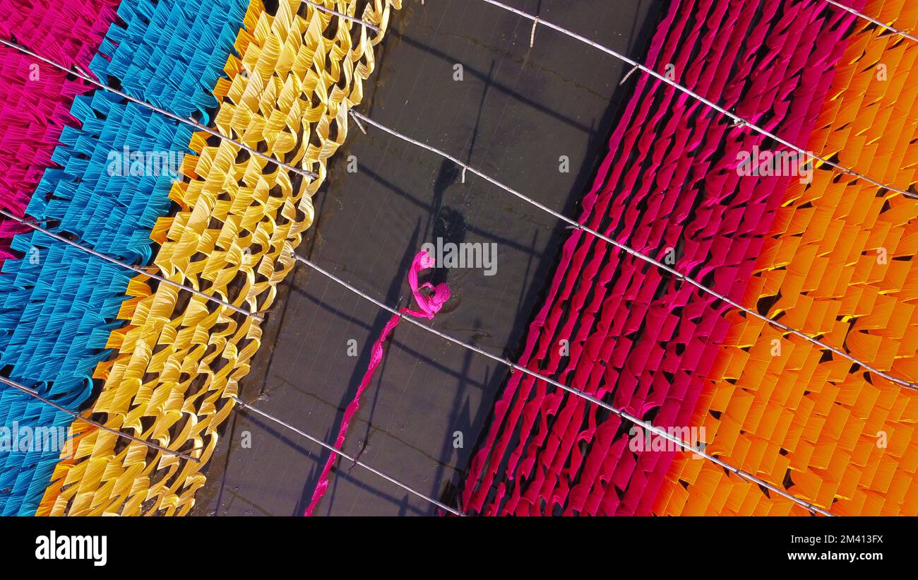 Narayanganj, Dhaka, Bangladesh. 18th Dec, 2022. Una scintillante rete di colori interconnessi produce un arcobaleno di colori mentre tessuti colorati vengono appesi ad asciugare sopra un campo allagato a Narayanganj, Bangladesh. I fili di ferro sono usati tra una struttura di bambù per generare le linee giganti di lavaggio per la parte finale del processo di tintura dopo che i panni sono fatti in t-shirt e giubbotti alla fabbrica dell'abbigliamento. Circa 4.000 pezzi di tessuto sono appesi ad asciugare qui ogni giorno. Il processo richiede solitamente cinque ore, con ogni set di 80 pezzi alla volta. Credit: ZUMA Press, Inc./Alamy Live News Foto Stock