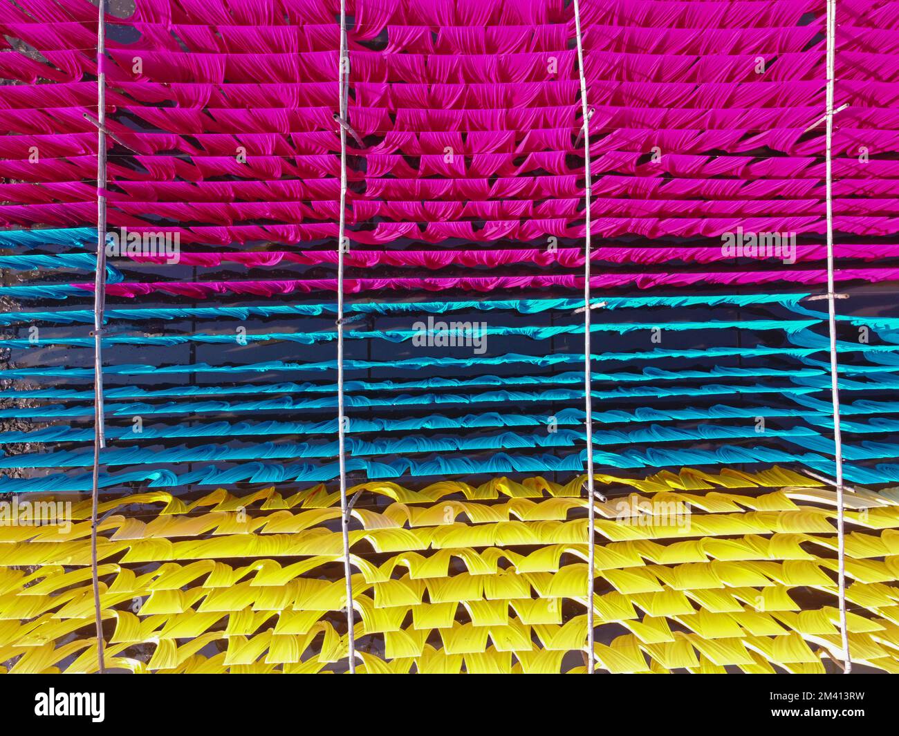 Narayanganj, Dhaka, Bangladesh. 18th Dec, 2022. Una scintillante rete di colori interconnessi produce un arcobaleno di colori mentre tessuti colorati vengono appesi ad asciugare sopra un campo allagato a Narayanganj, Bangladesh. I fili di ferro sono usati tra una struttura di bambù per generare le linee giganti di lavaggio per la parte finale del processo di tintura dopo che i panni sono fatti in t-shirt e giubbotti alla fabbrica dell'abbigliamento. Circa 4.000 pezzi di tessuto sono appesi ad asciugare qui ogni giorno. Il processo richiede solitamente cinque ore, con ogni set di 80 pezzi alla volta. Credit: ZUMA Press, Inc./Alamy Live News Foto Stock