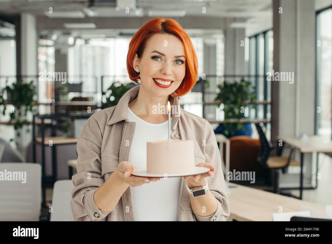 Felice donna d'affari che tiene la torta di compleanno mentre fa la festa di sorpresa con i suoi colleghi in ufficio. Foto Stock