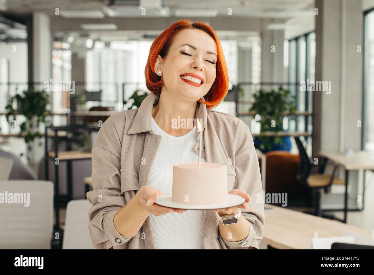 Felice donna d'affari che tiene la torta di compleanno mentre fa la festa di sorpresa con i suoi colleghi in ufficio. Foto Stock