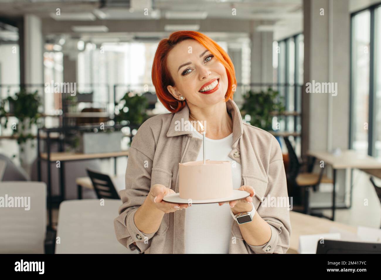 Felice donna d'affari che tiene la torta di compleanno mentre fa la festa di sorpresa con i suoi colleghi in ufficio. Foto Stock