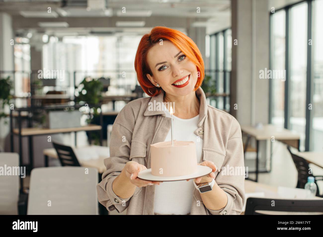 Felice donna d'affari che tiene la torta di compleanno mentre fa la festa di sorpresa con i suoi colleghi in ufficio. Foto Stock
