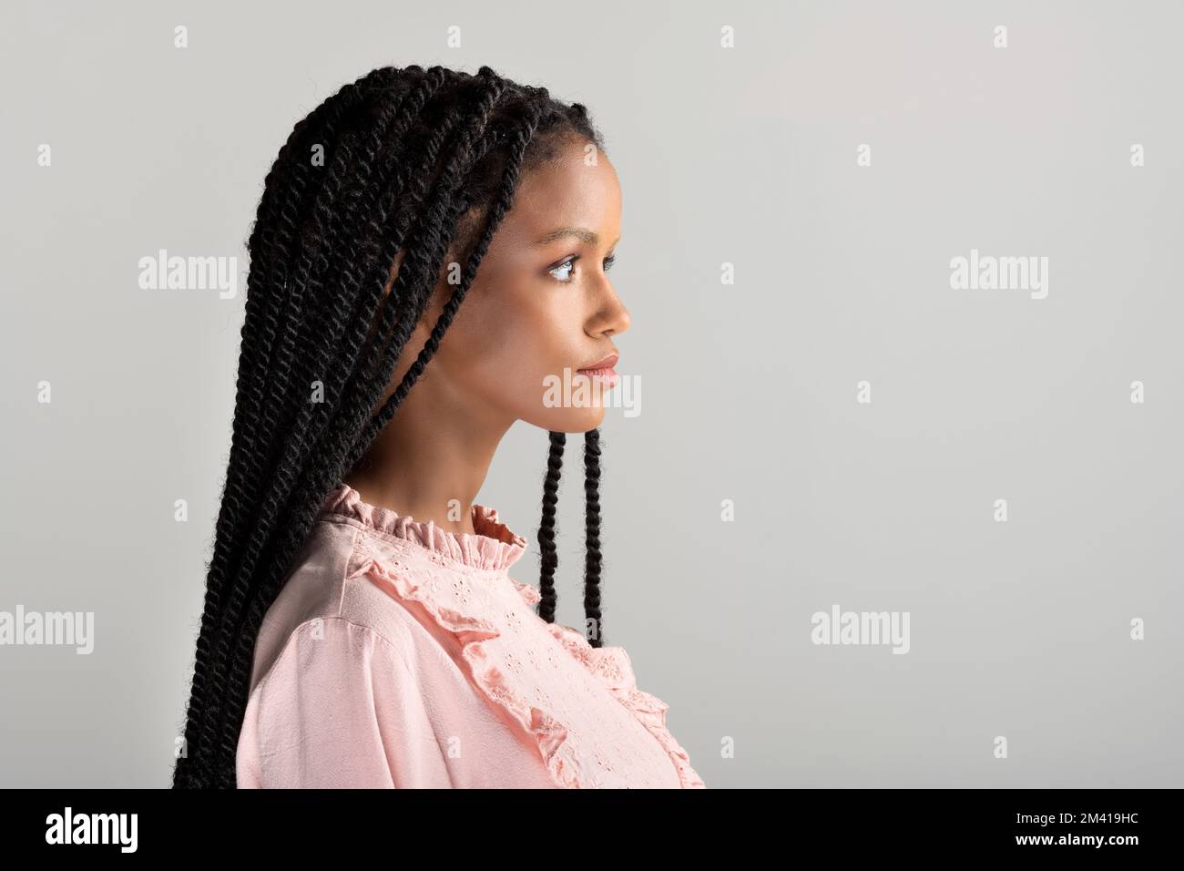 Vista laterale della giovane ragazza afro-americana in blusa rosa vintage con lunghi capelli intrecciati che guardano lontano da sfondo grigio Foto Stock