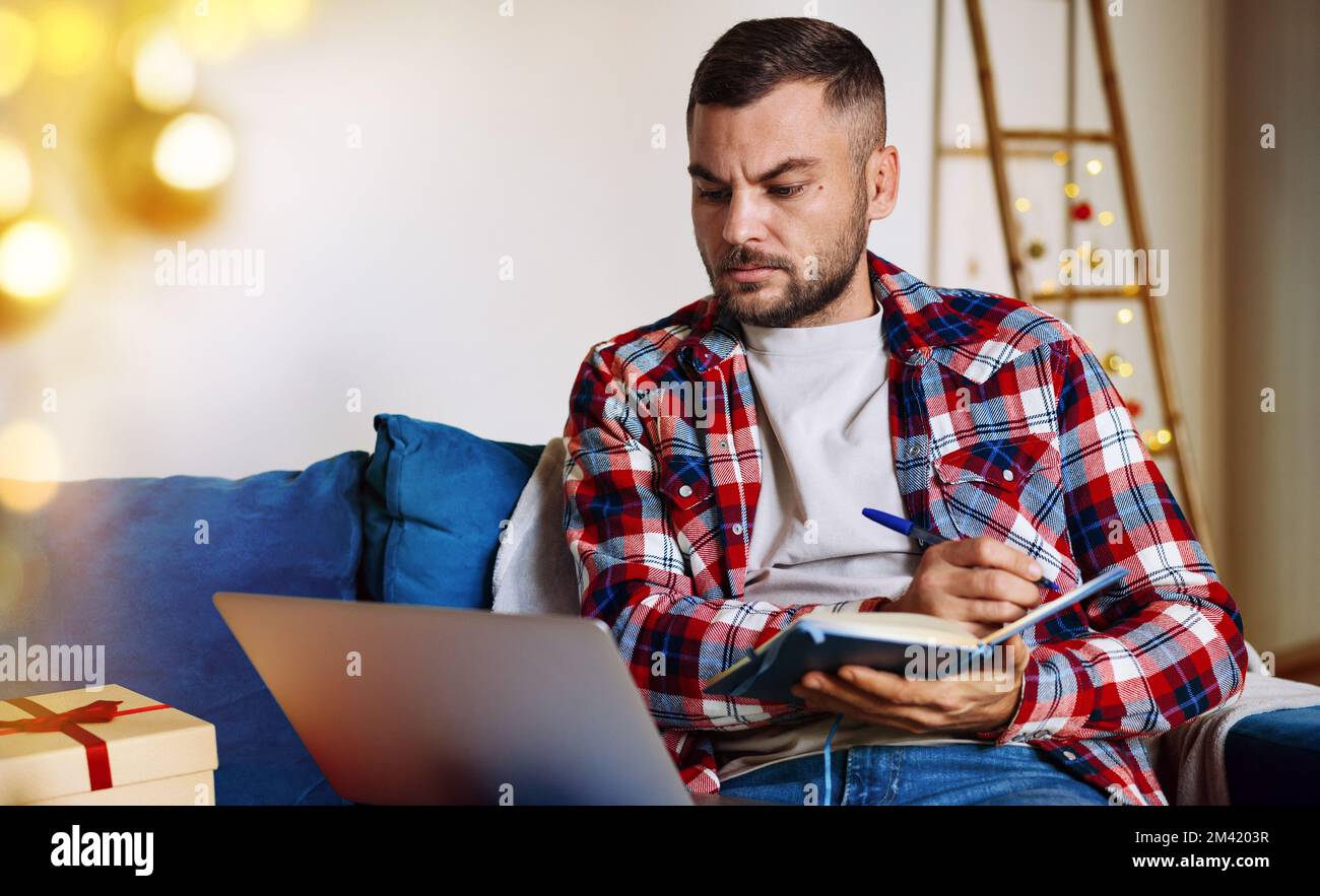 Uomo che lavora da solo a casa durante le vacanze di Natale utilizzando un notebook e scrivendo appunti nel suo notebook. Foto Stock