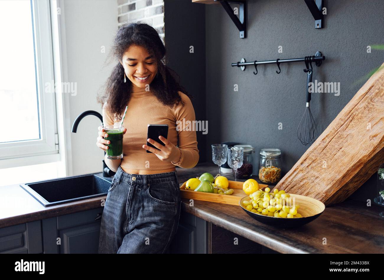 Donna afro che tiene una bevanda di verdure. Giovane ragazza afro-americana bere succo verde con cannuccia di bambù riutilizzabile e leggere notizie sul telefono cellulare Foto Stock