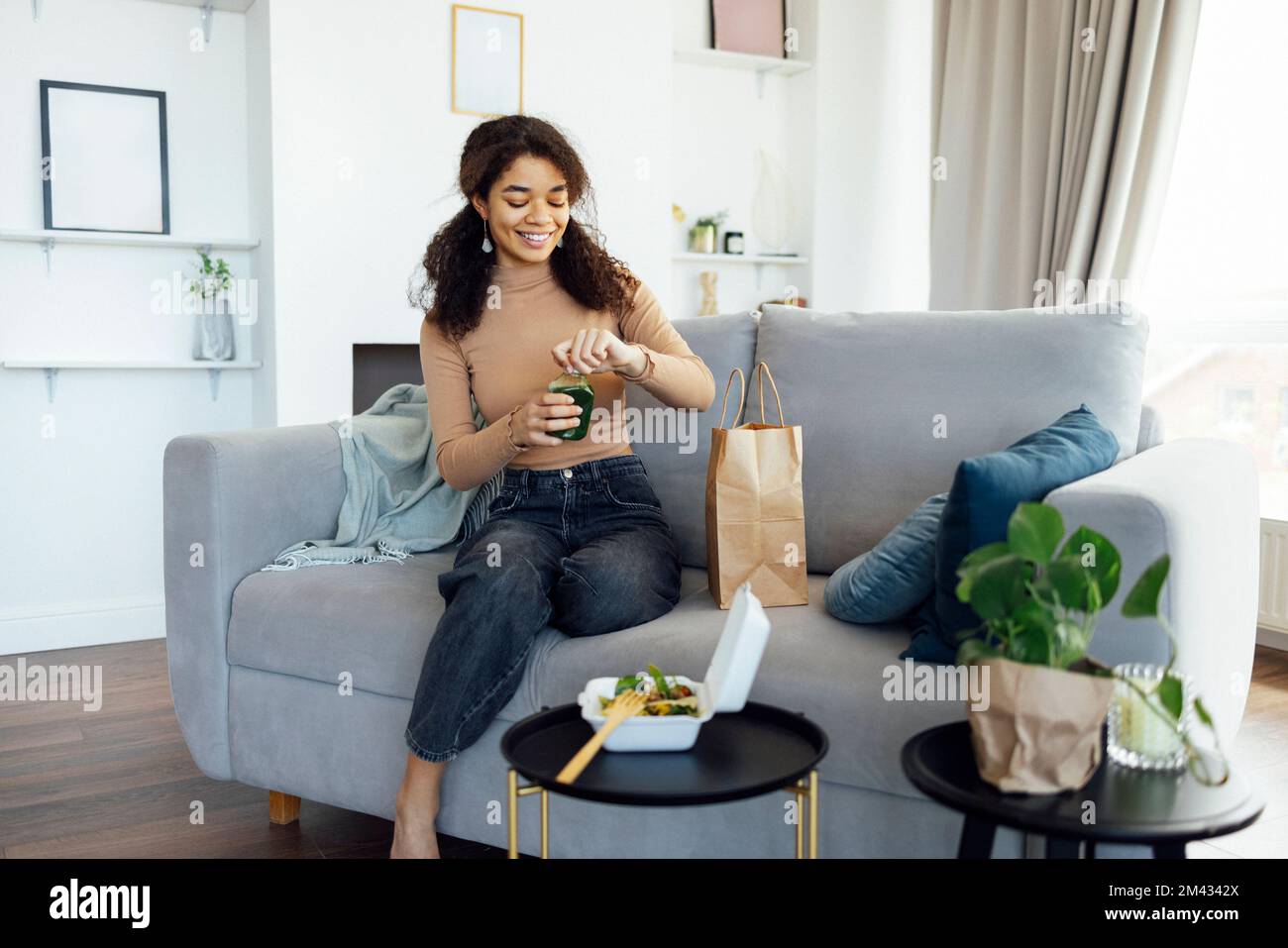 Donna afro-americana soddisfatta che mangia sana insalata di pasto equilibrata e verde fresco o frullato consegnato dal servizio di consegna del cibo. La donna a casa gode Foto Stock