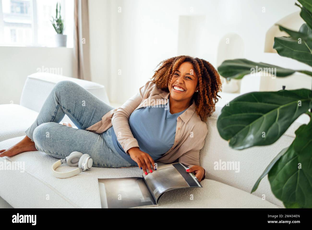 Foto di bella felice giovane donna africana seduto sul divano in casa durante la lettura di riviste o libri Foto Stock