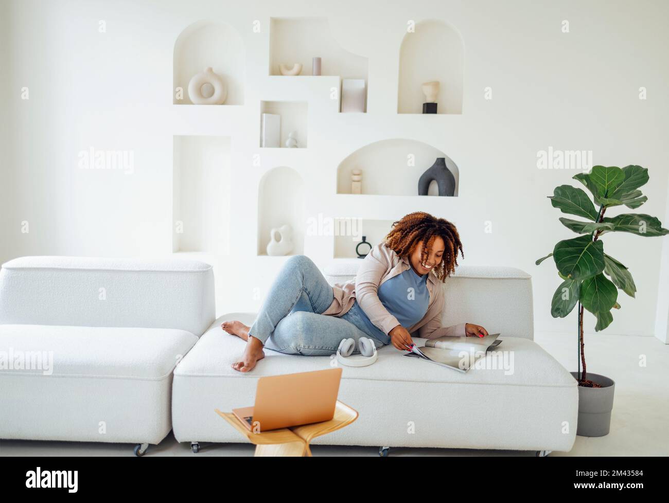 Foto di bella felice giovane donna africana seduto sul divano in casa durante la lettura di riviste o libri Foto Stock