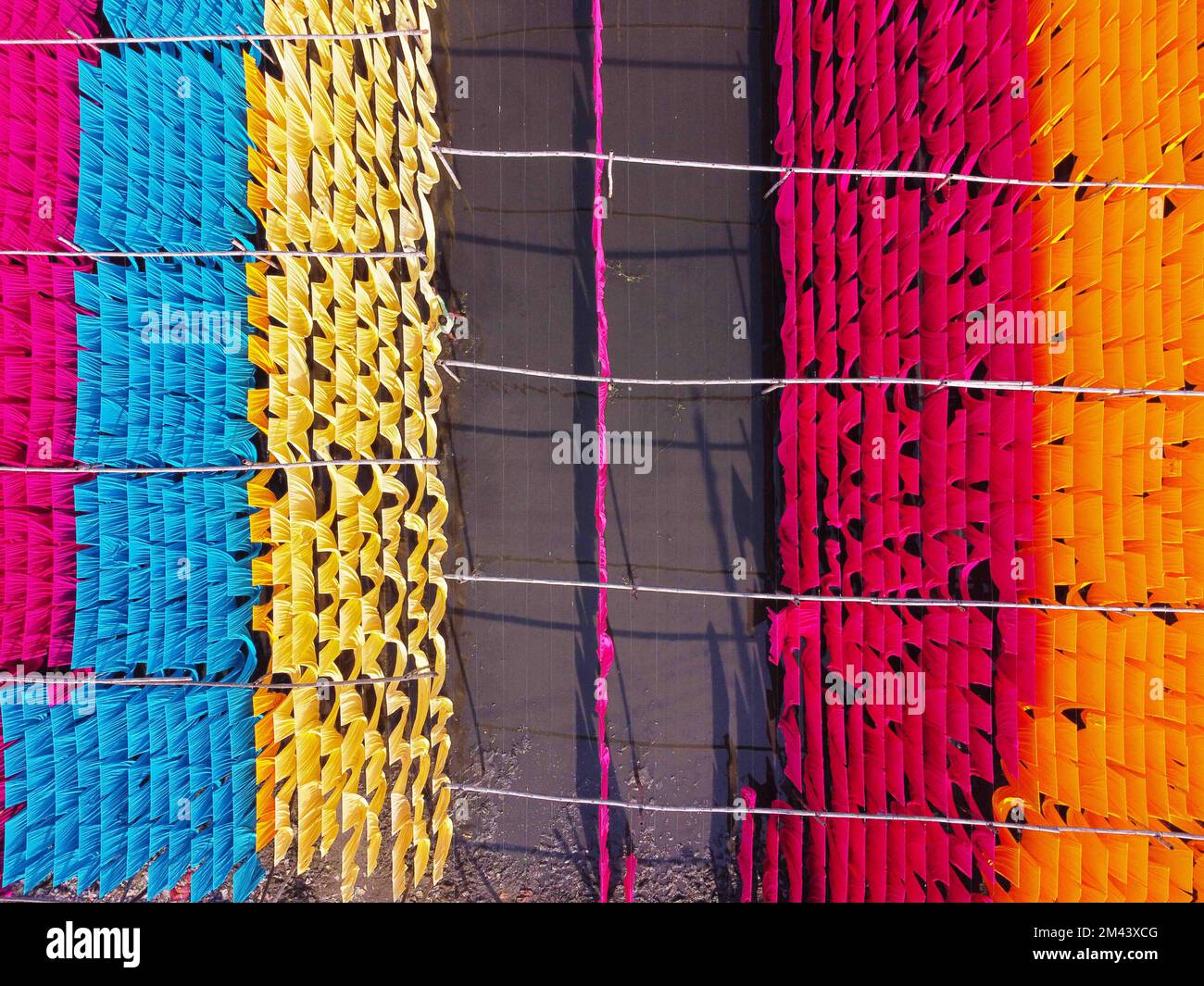 Narayanganj, Dhaka, Bangladesh. 18th Dec, 2022. Una scintillante rete di colori interconnessi produce un arcobaleno di colori mentre tessuti colorati vengono appesi ad asciugare sopra un campo allagato a Narayanganj, Bangladesh. I fili di ferro sono usati tra una struttura di bambù per generare le linee giganti di lavaggio per la parte finale del processo di tintura dopo che i panni sono fatti in t-shirt e giubbotti alla fabbrica dell'abbigliamento. Circa 4.000 pezzi di tessuto sono appesi ad asciugare qui ogni giorno. Il processo richiede solitamente cinque ore, con ogni set di 80 pezzi alla volta. Dopo che i vestiti sono tinti usando le macchine nella fabbrica, Foto Stock