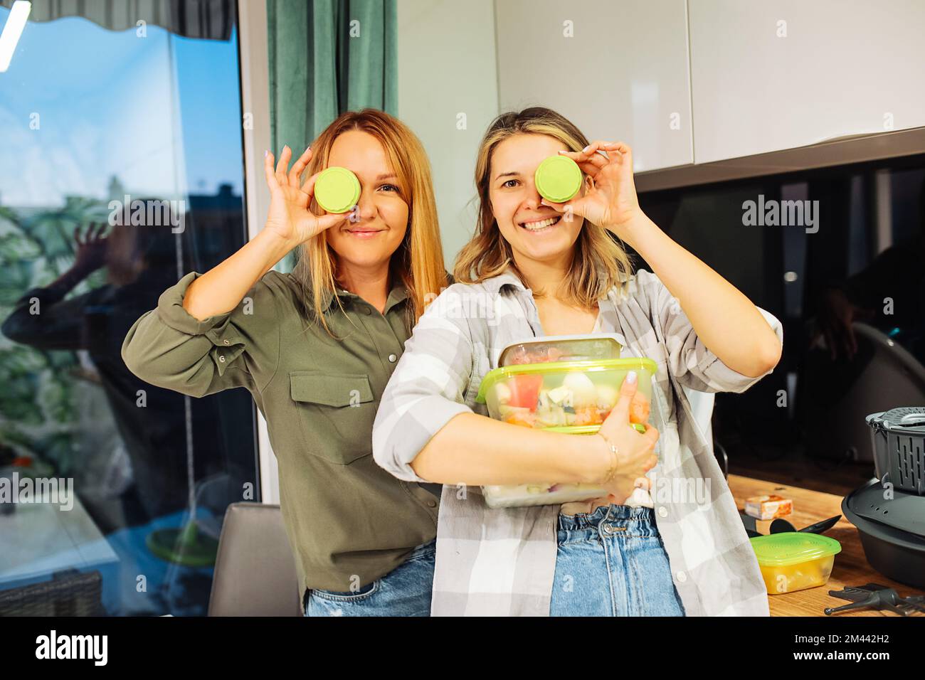 Due donne felici tengono i coperchi verdi davanti agli occhi. Amici che cucinano insieme in cucina. Alimenti in contenitori di plastica. Foto Stock
