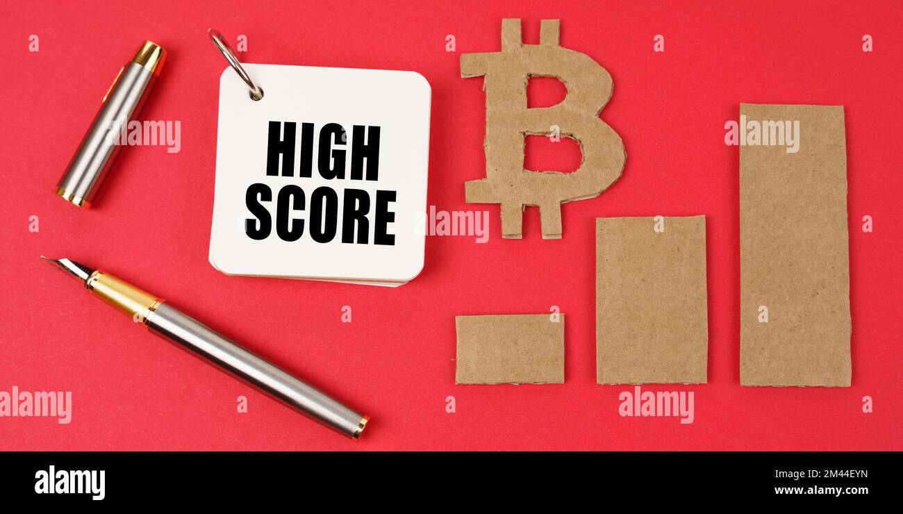 Business e bitcoin concetto. Su una superficie rossa si trovano un simbolo di bitcoin, un grafico, una penna e un blocco note con l'iscrizione - punteggio alto Foto Stock