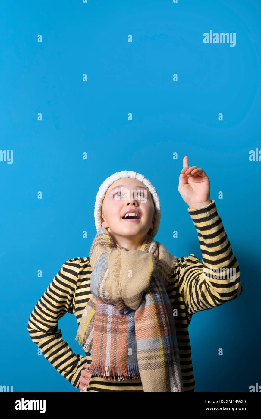 Una ragazza adolescente nei vestiti di inverno punta in su alla bandiera con il suo dito indice allo spazio per il testo. Ritratto verticale di una bella ragazza su un blu Foto Stock