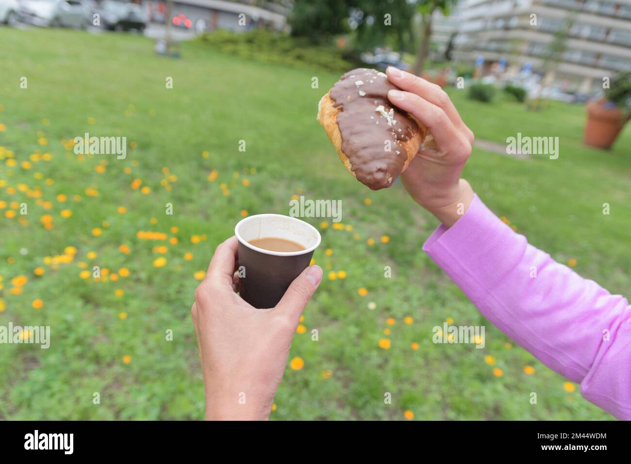 Stile di vita della città, caffè e croissant al cioccolato in città in un prato parco con fiori. In primo piano con le mani femminili Foto Stock