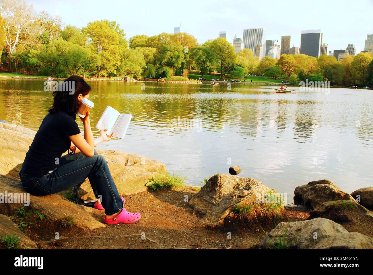 Una giovane donna legge un libro con una tazza di caffè ai margini del lago Central Park con vista sullo skyline di New York Foto Stock