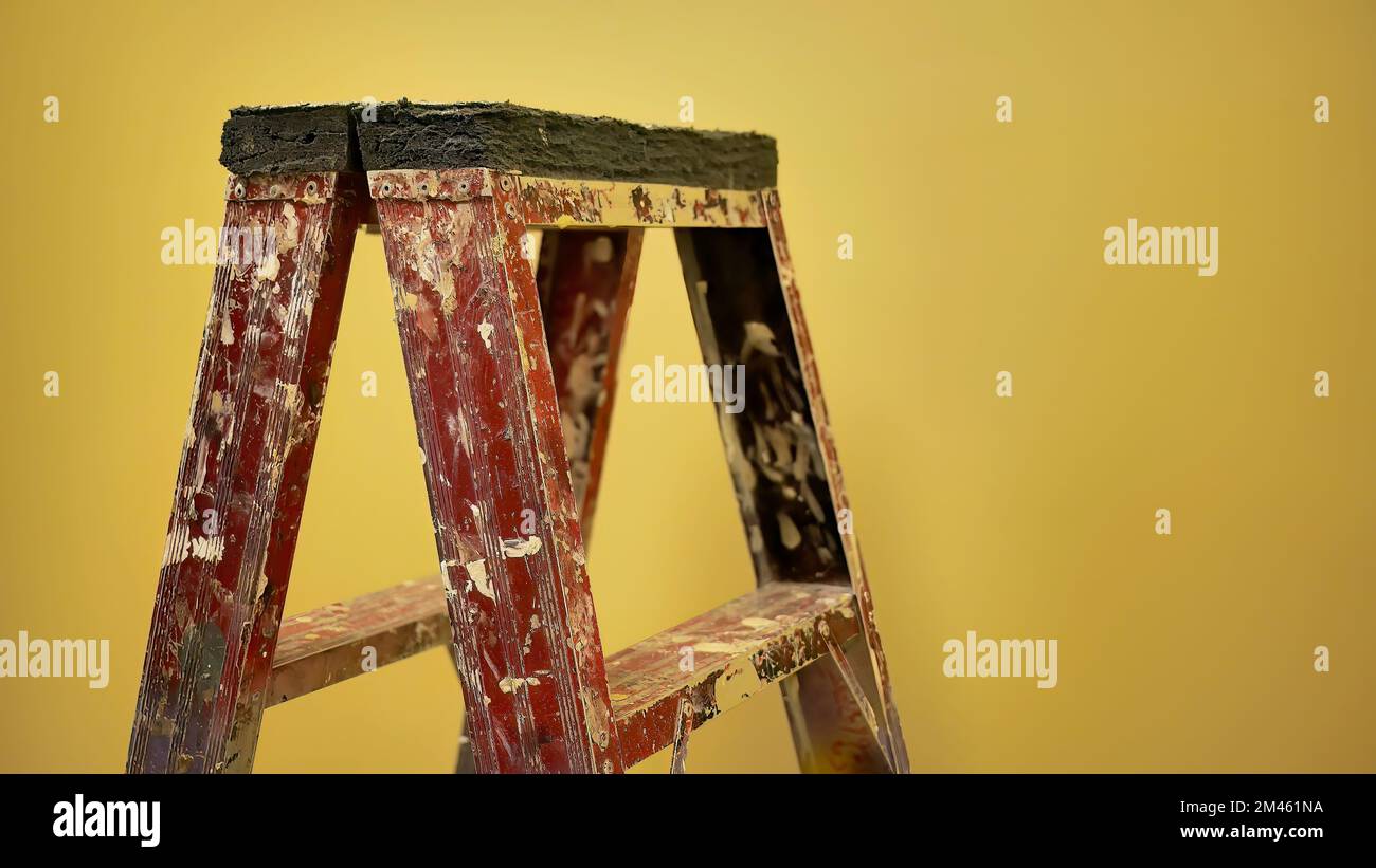 Vecchia scala per pittori di costruzione con spruzzi dipinti che si trovano di fronte alla parete gialla sotto una calda illuminazione bianca Foto Stock