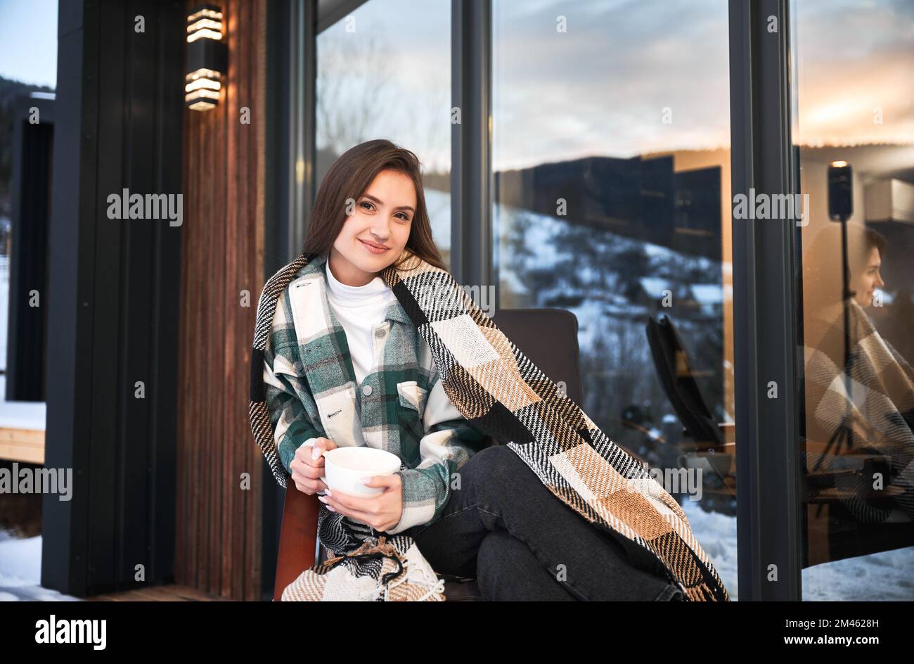 Giovane donna che riposa sulla terrazza di una moderna casa di fienile in montagna. Felice femmina turista che tiene una tazza di tè, godendo la vacanza invernale in un nuovo cottage. Foto Stock