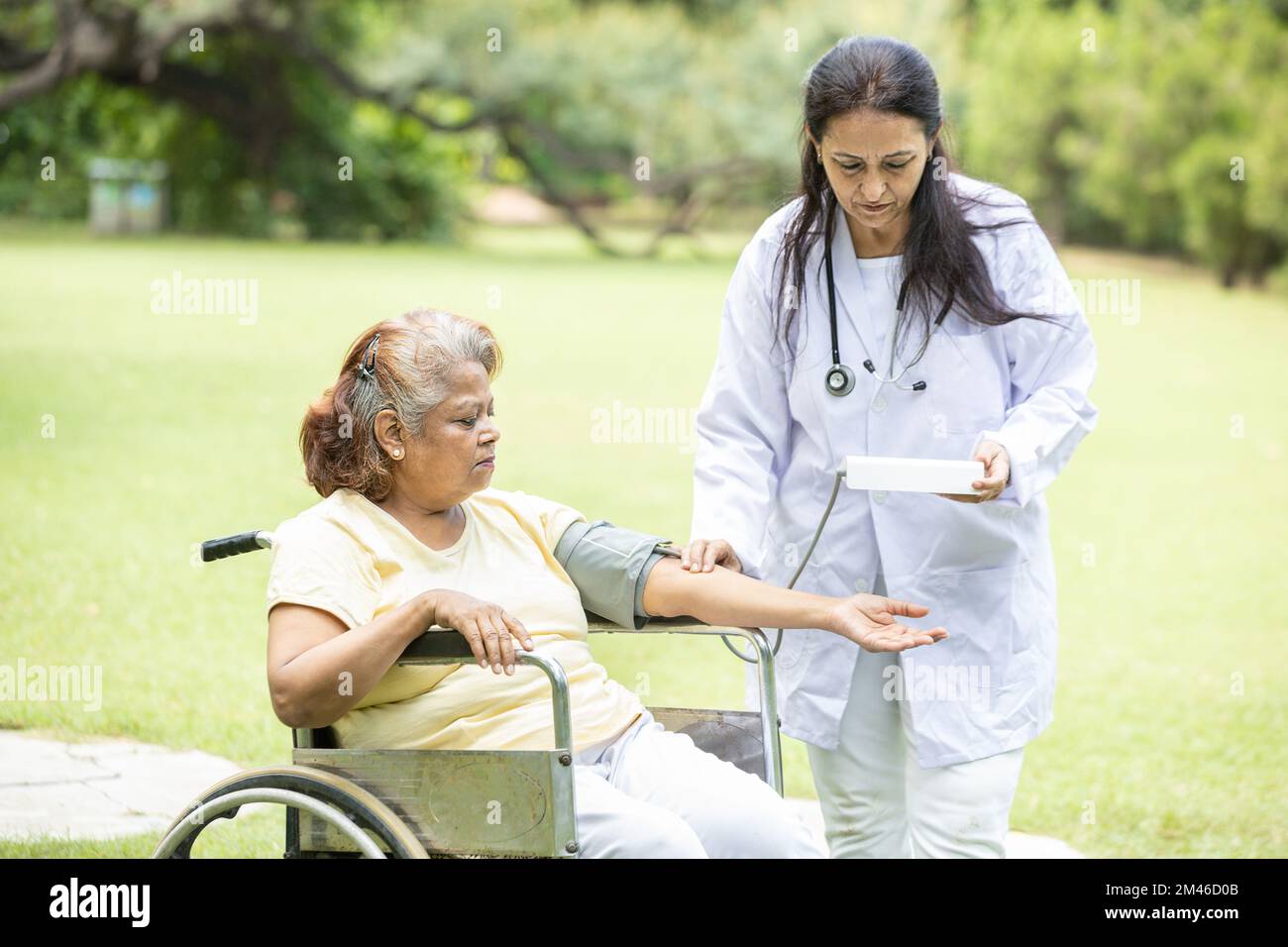 Il medico indiano controlla la pressione sanguigna della paziente anziana in una sedia a rotelle all'aperto nel parco. Foto Stock