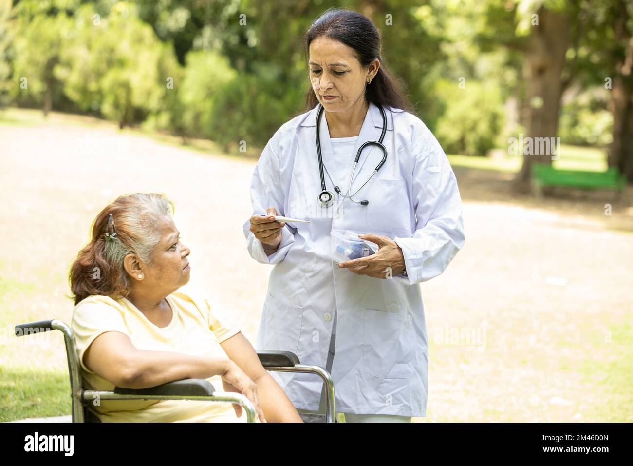 Il medico indiano o l'infermiere misurano la temperatura corporea con il termometro paziente anziano in una sedia a rotelle all'aperto nel parco. Foto Stock
