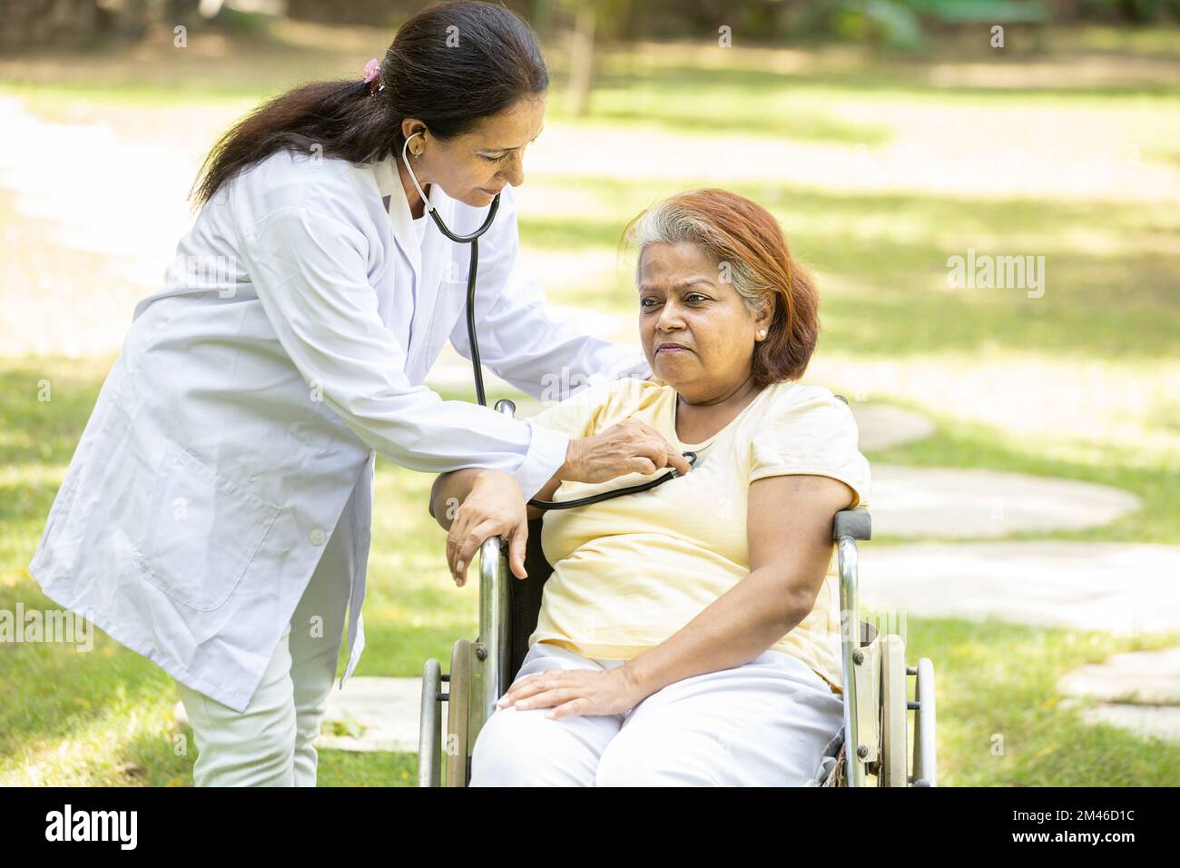 Il medico indiano controlla il battito cardiaco di una donna anziana in una sedia a rotelle all'aperto nel parco. Foto Stock