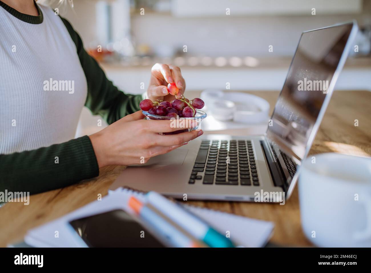 Vista laterale della donna che tiene il recipiente con uva sopra la scrivania con computer, diario e smartphone. Concetto di equilibrio tra vita lavorativa e vita privata. Foto Stock