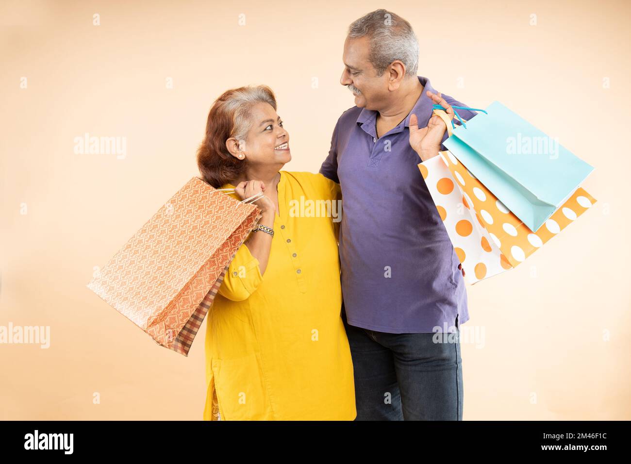 Felice coppia indiana senior in possesso di borse shopping guardando l'un l'altro con isolato su sfondo beige. la vita di pensione, le vendite e le offerte di e-commercio Foto Stock