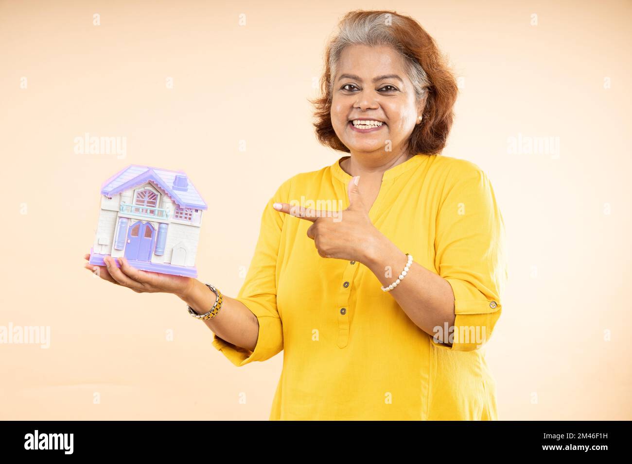 Donna indiana anziana felice con una casa giocattolo in mano isolata su sfondo beige, prestito casa, proprietà in affitto, la vita di pensione. Immobiliare e alloggio Foto Stock