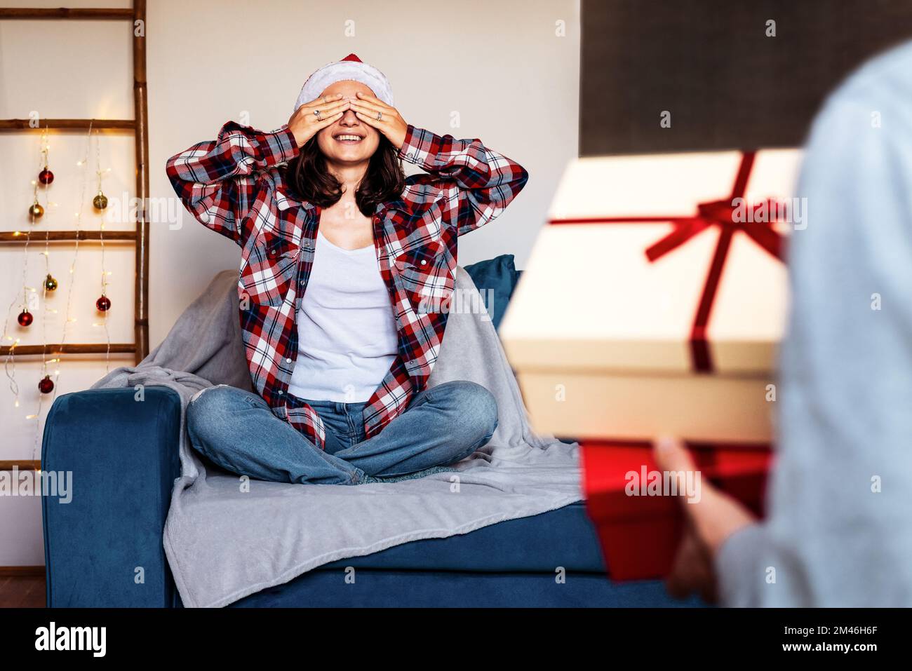 Giovane donna che indossa il cappello di Santa seduta sul divano con gli occhi chiusi con le mani mentre il suo ragazzo che porta le scatole del regalo di Natale. Foto Stock