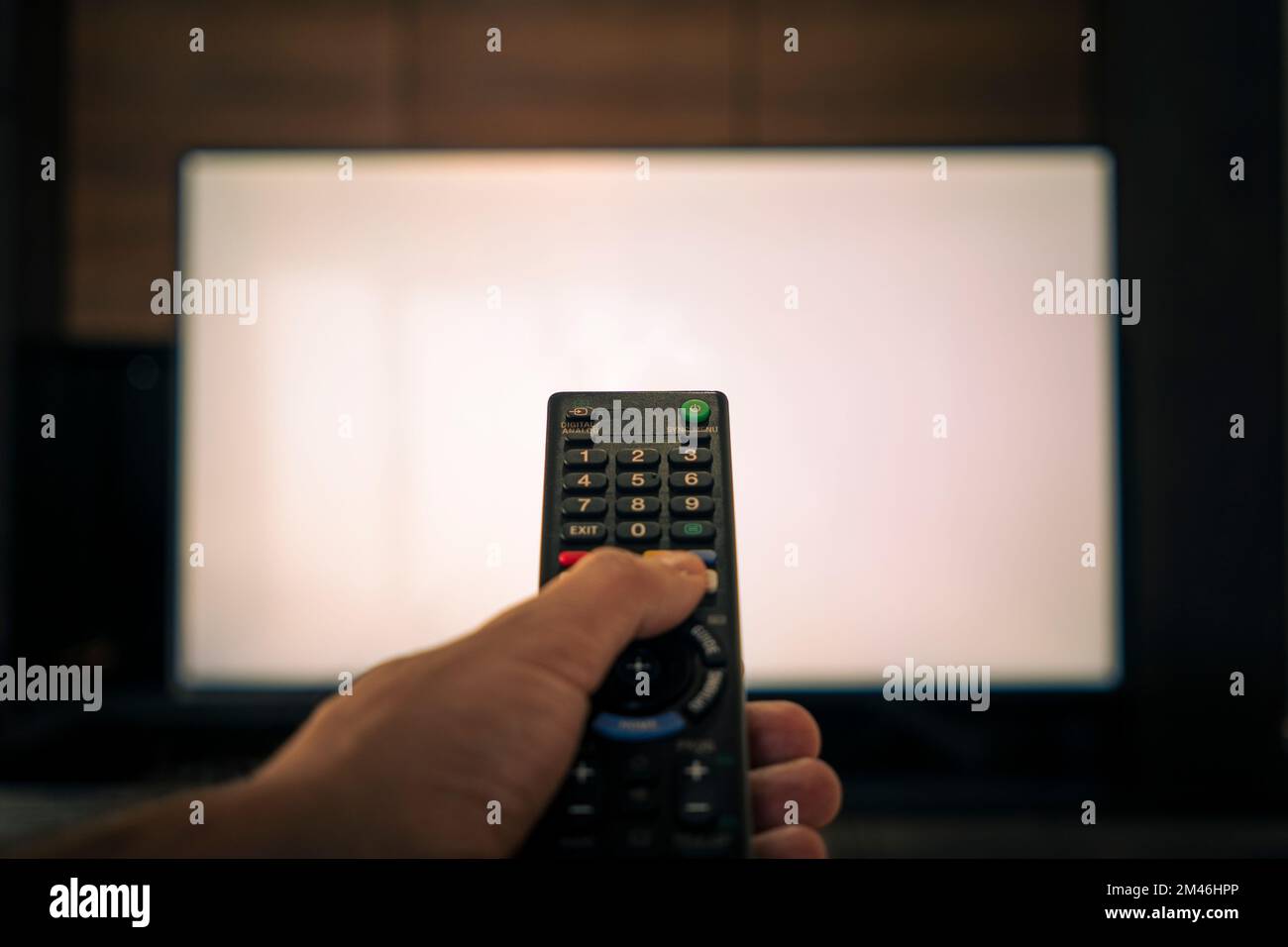 telecomando sfocato per il televisore. La mano di un uomo tiene il telecomando davanti a un grande schermo TV bianco. Foto Stock