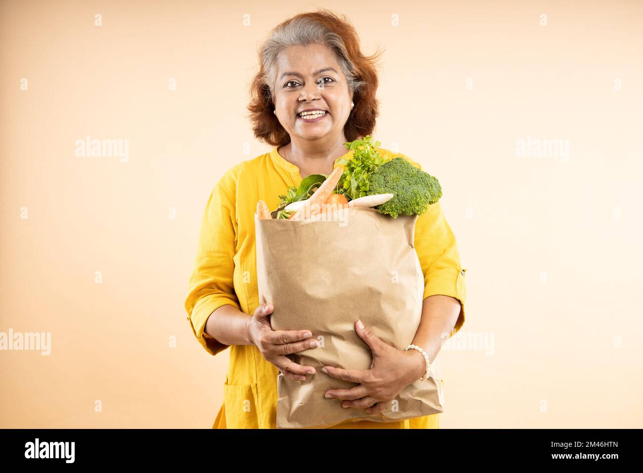 Donna indiana anziana felice tenere sacchetti di carta della drogheria con cibo isolato su sfondo beige. foto da studio. Foto Stock