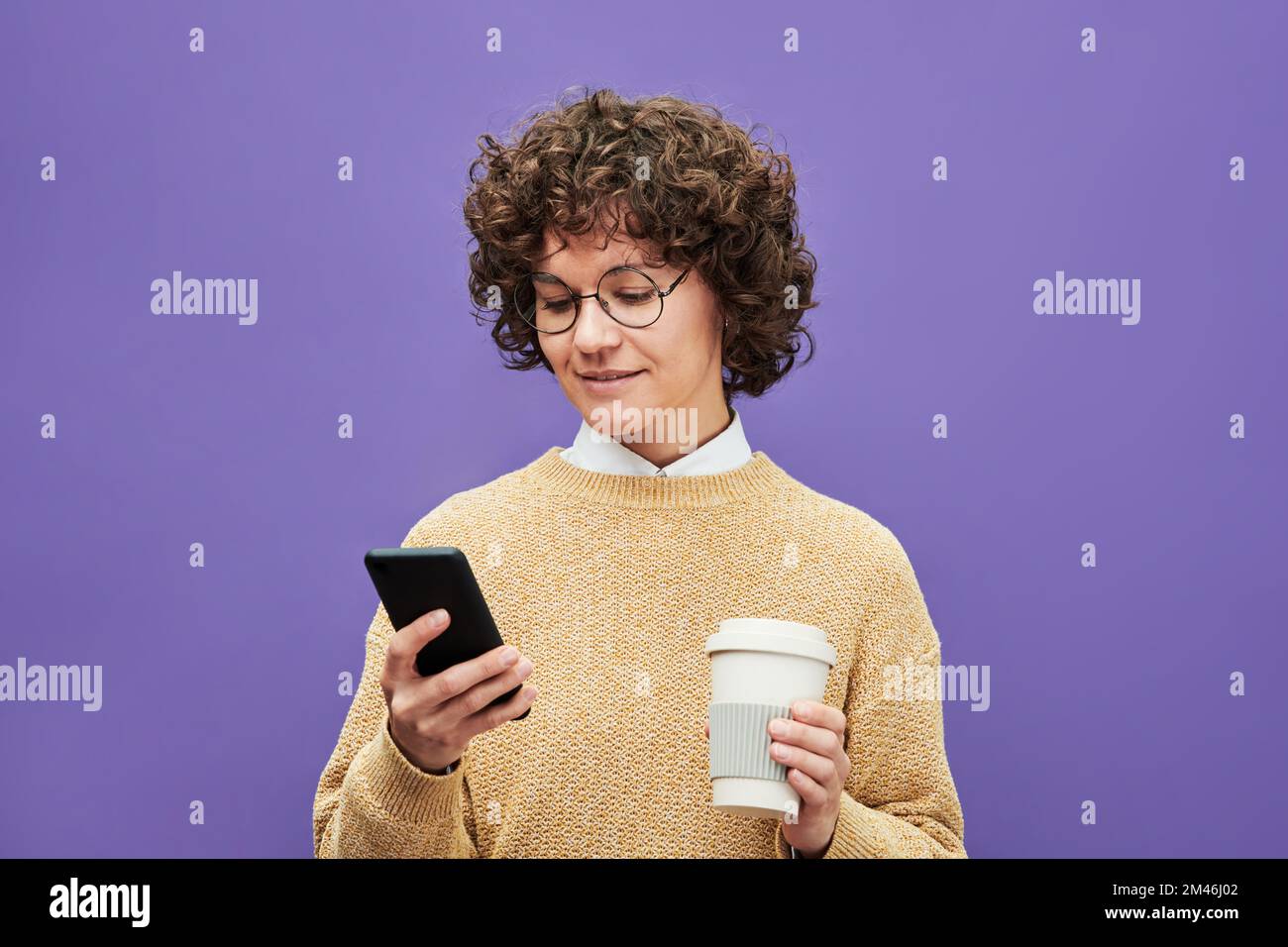 Giovane donna brunetta in un pullover casual guardando attraverso le notizie online in smartphone e avendo una tazza di caffè su sfondo viola Foto Stock