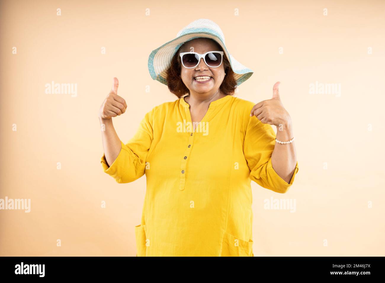 Donna indiana anziana felice che indossa cappello e occhiali da sole fare pollici con entrambe le mani in piedi isolato su sfondo beige studio, la vita di pensionamento, holida Foto Stock