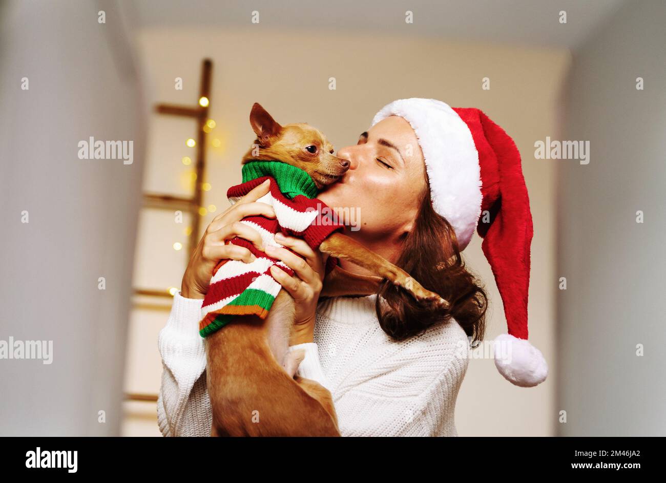 Donna che indossa il cappello di Santa che tiene il cane piccolo nelle sue mani e baciando disimballare il concetto di regali di Natale. Foto Stock