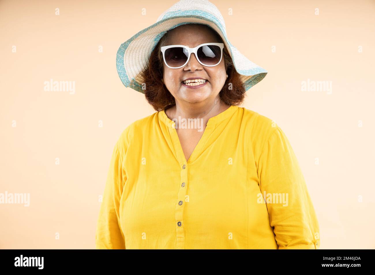 Donna indiana anziana felice che indossa cappello e occhiali da sole in piedi isolato su sfondo beige studio, la vita di pensionamento, vacanze e viaggi. primo piano Foto Stock