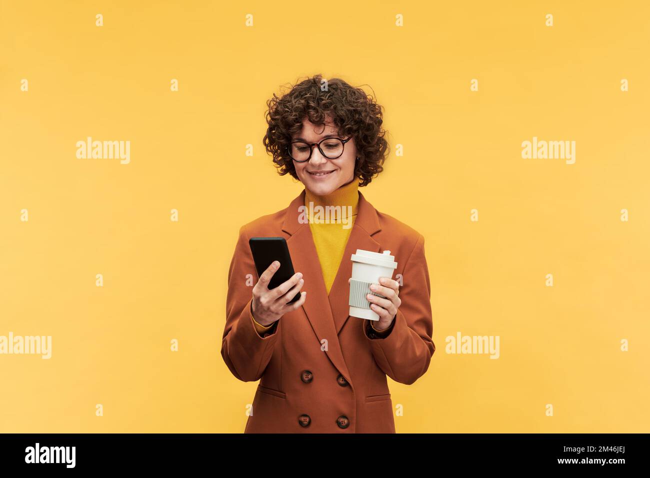Giovane donna brunetta sorridente in formalwear che ha una tazza di caffè e guardando attraverso le foto online o le notizie in telefono cellulare Foto Stock