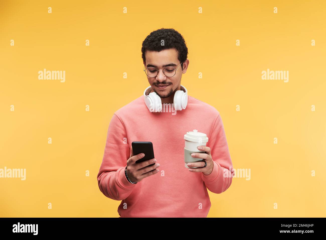 Giovane uomo multietnico con cuffie bianche intorno al collo utilizzando lo smartphone e una tazza di caffè su sfondo giallo Foto Stock
