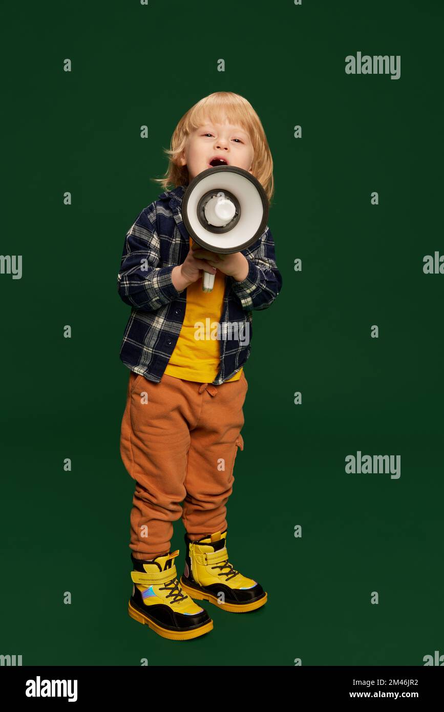 Ritratto di ragazzino, bambino in posa con megafone su sfondo verde studio. Vendite, notizie. Concetto di infanzia, emozioni Foto Stock