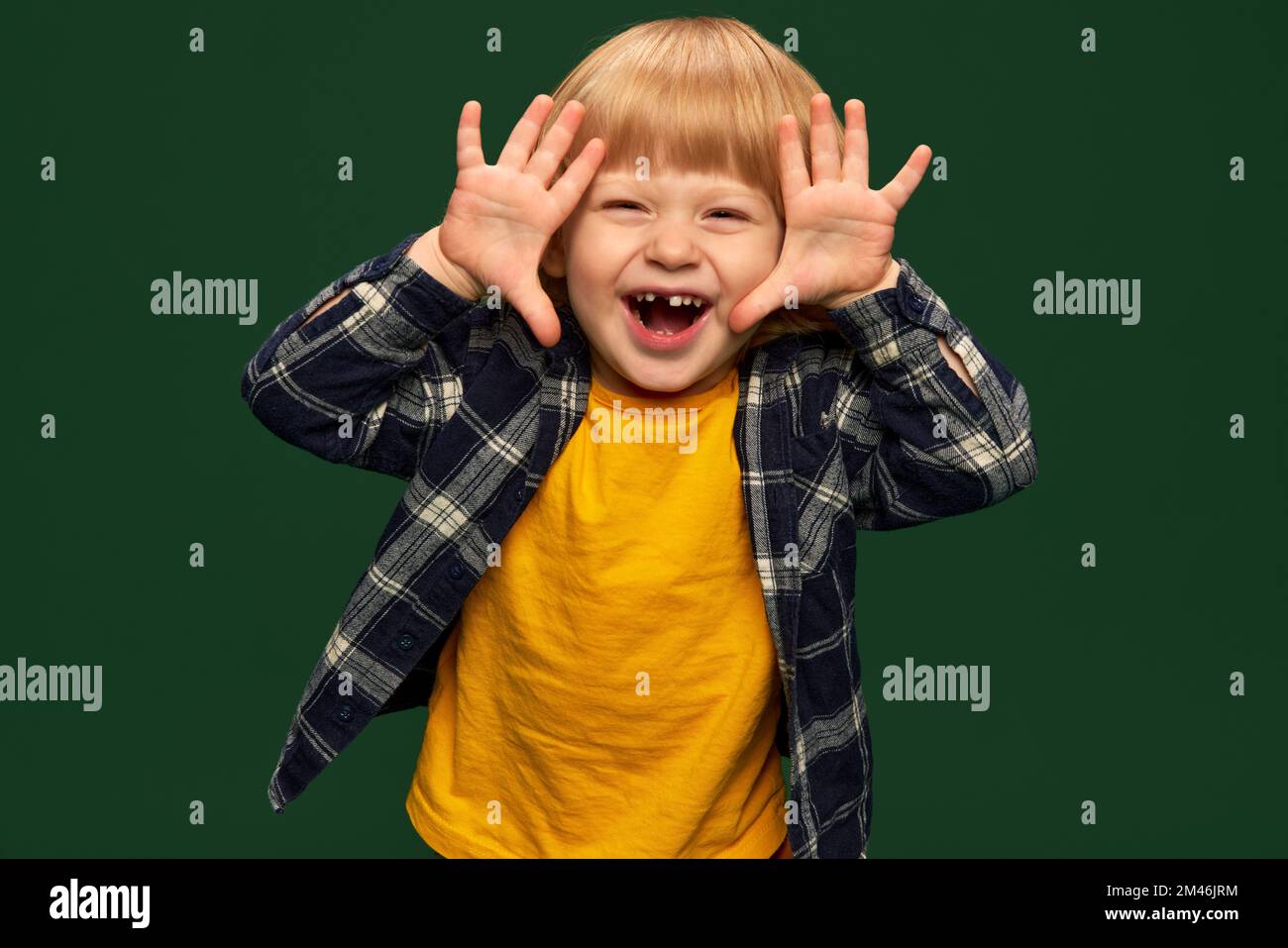 Ritratto di carino ragazzino, bambino in posa, sorridente su sfondo verde studio. Giocare a nascondere e cercare. Concetto di infanzia, emozioni Foto Stock