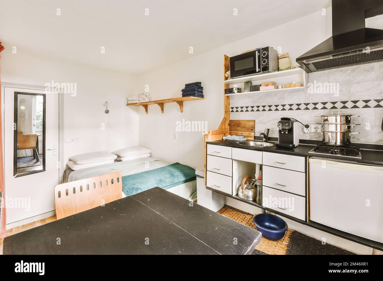 una zona cucina in un piccolo appartamento con armadi bianchi e ripiani neri alle pareti, una finestra aperta sulla camera è Foto Stock