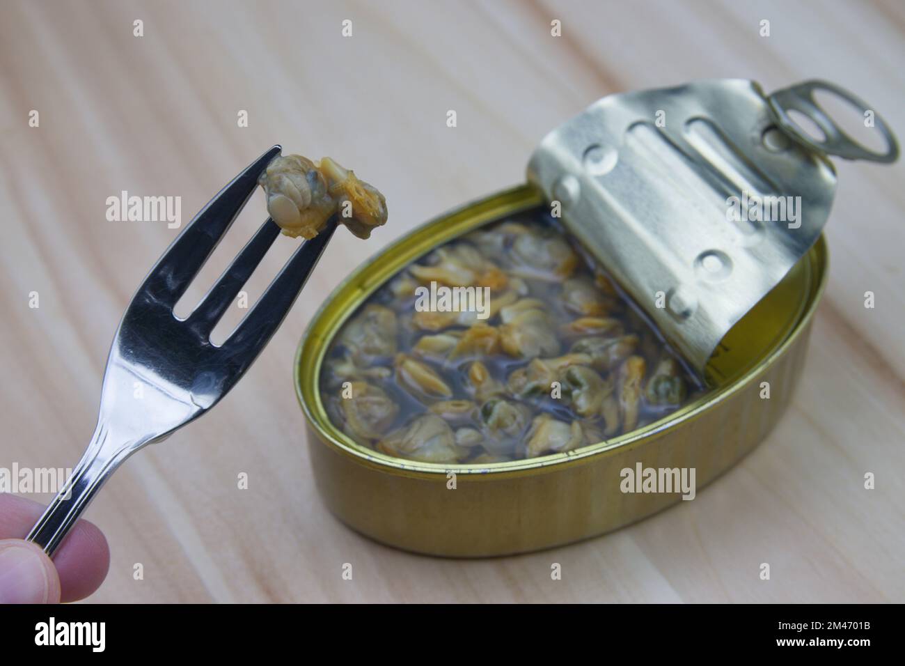 Primo piano di una lattina di cockles accanto a qualcuno che tiene una forchetta con una cockle su un tavolo di legno Foto Stock