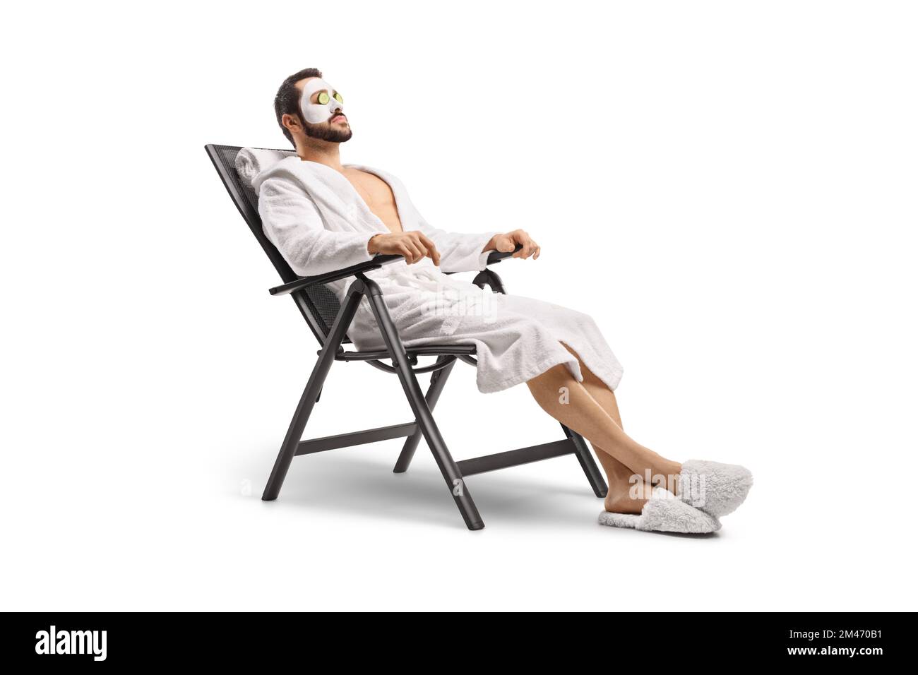 Uomo in accappatoio con maschera facciale e cetriolo sugli occhi seduti su una sedia isolata su sfondo bianco Foto Stock